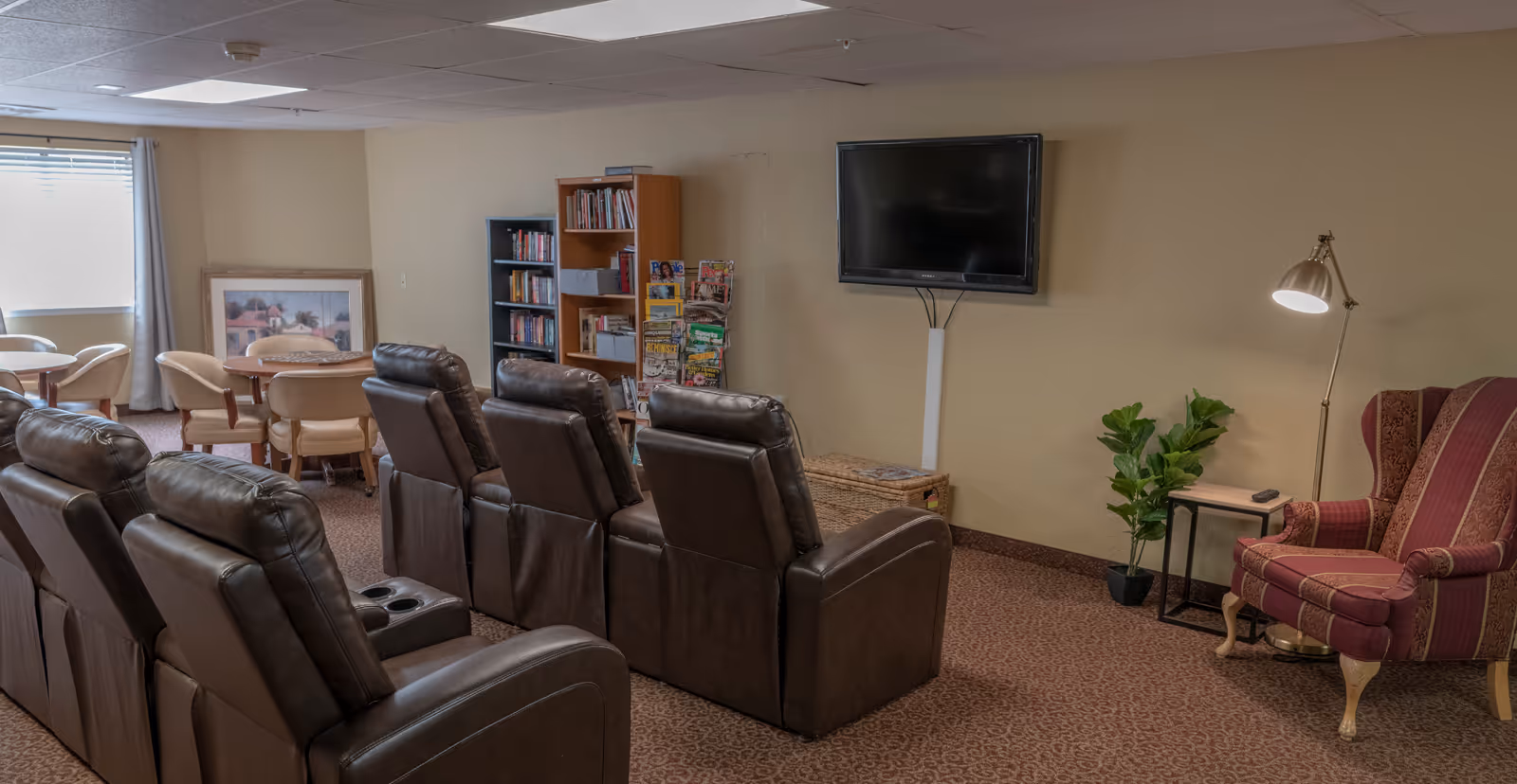 A senior living common room with rows of leather recliners facing a wall-mounted TV, a bookshelf and tables with chairs in the background.