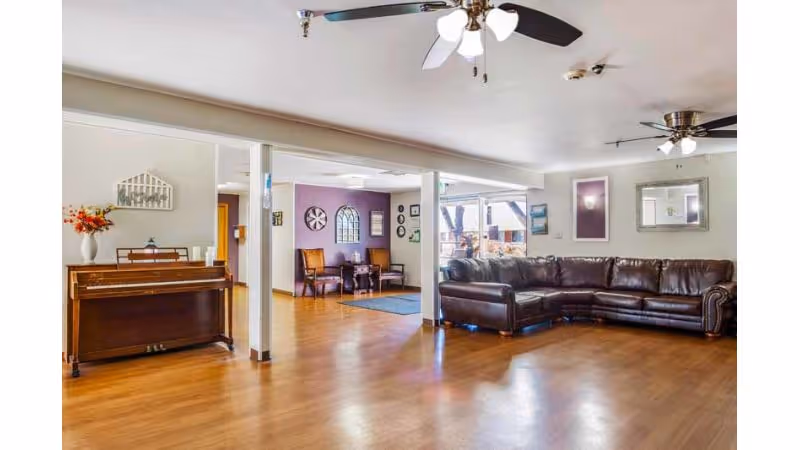 A spacious living room area with wooden flooring, featuring a large brown leather sectional sofa on the right side. Two ceiling fans with lights are mounted on the white ceiling. On the left side, there is an upright piano with a vase of flowers on top. In the background, a small seating area with two chairs and a table is visible against a purple accent wall decorated with clocks and framed pictures. Large windows allow natural light to fill the room.