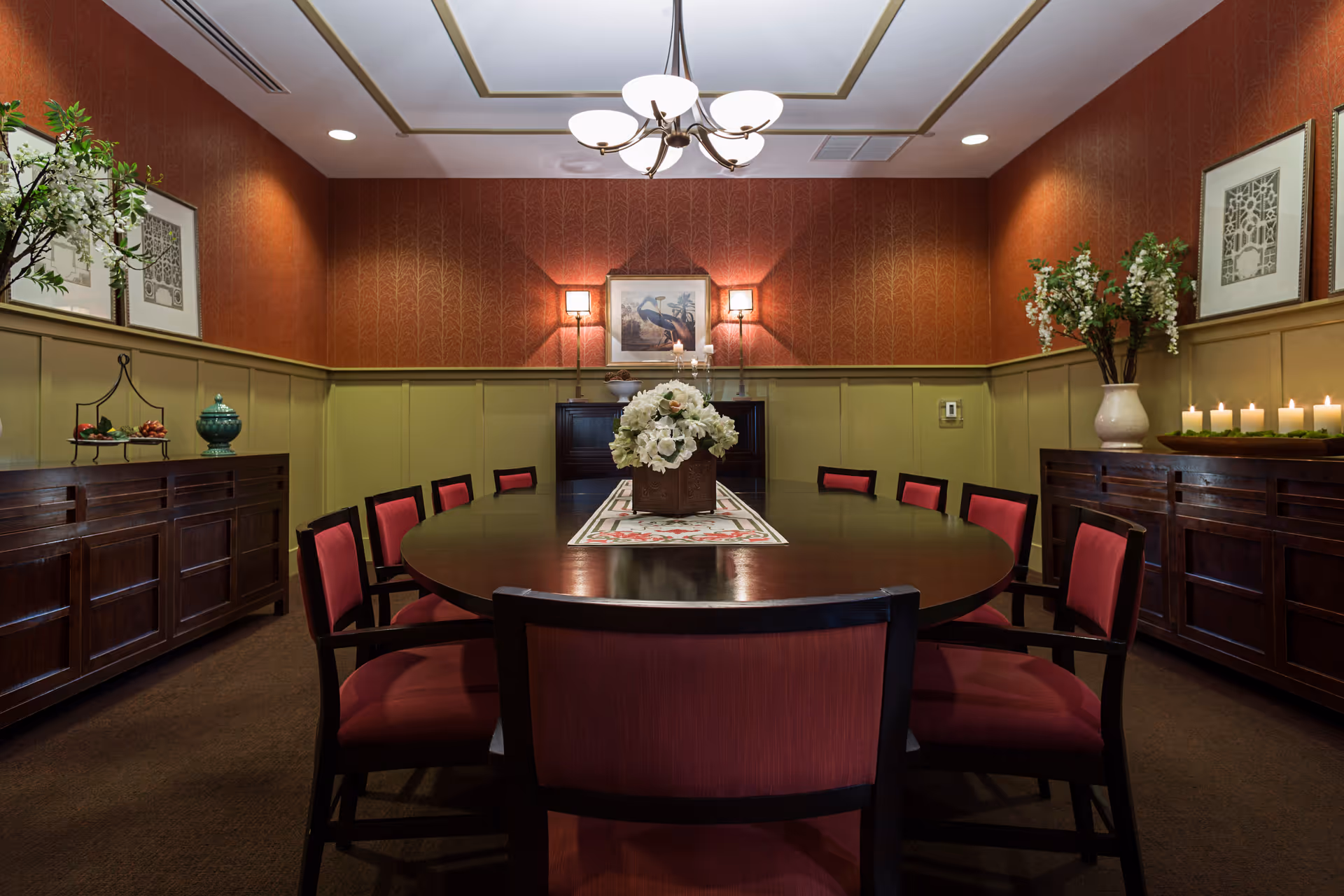 A formal dining room with a large oval wooden table surrounded by red cushioned chairs. The room features warm-toned walls with a chair rail, decorative framed artwork, a chandelier overhead, and floral arrangements on sideboards and the table. Candles are lit on one sideboard, creating a cozy ambiance.