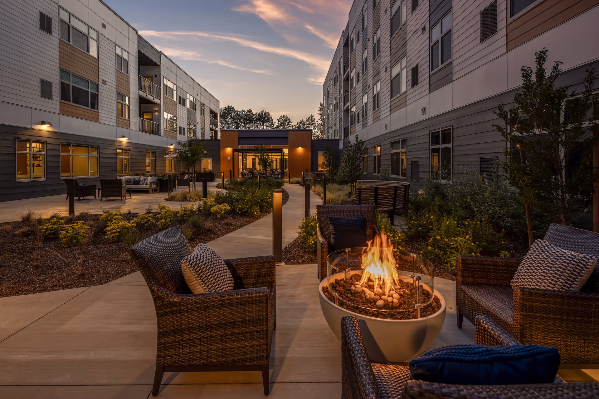 Outdoor courtyard area at dusk between two multi-story residential buildings with seating arrangements including wicker chairs with cushions around a lit fire pit, landscaped garden beds, and a paved walkway leading to a building entrance.