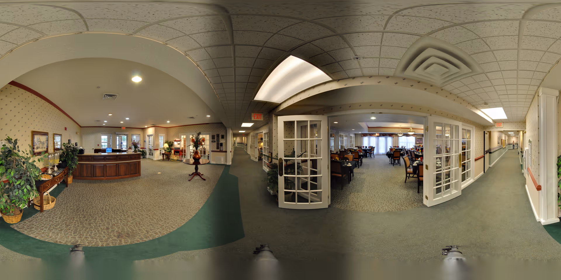 Panoramic view of the interior of Smith Farms Manor showing a reception desk area with plants and decorations on the left, a hallway extending to the right, and a dining room with tables and chairs visible through glass double doors in the center.