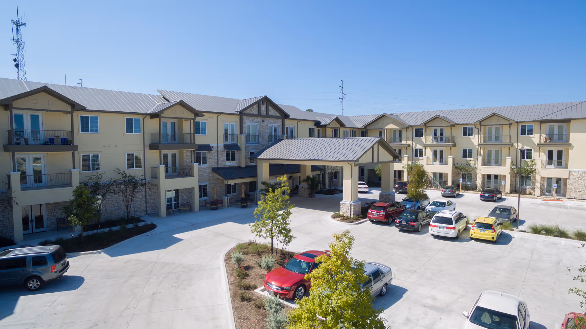 Front exterior of a three-story senior living building with a covered entrance, balconies, landscaping, and a parking lot with cars.