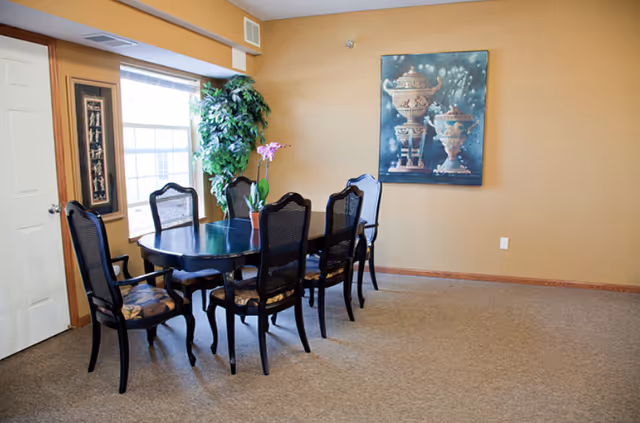 Dining room with a dark wood table and six chairs, a potted plant by a window, and artwork on a tan wall.