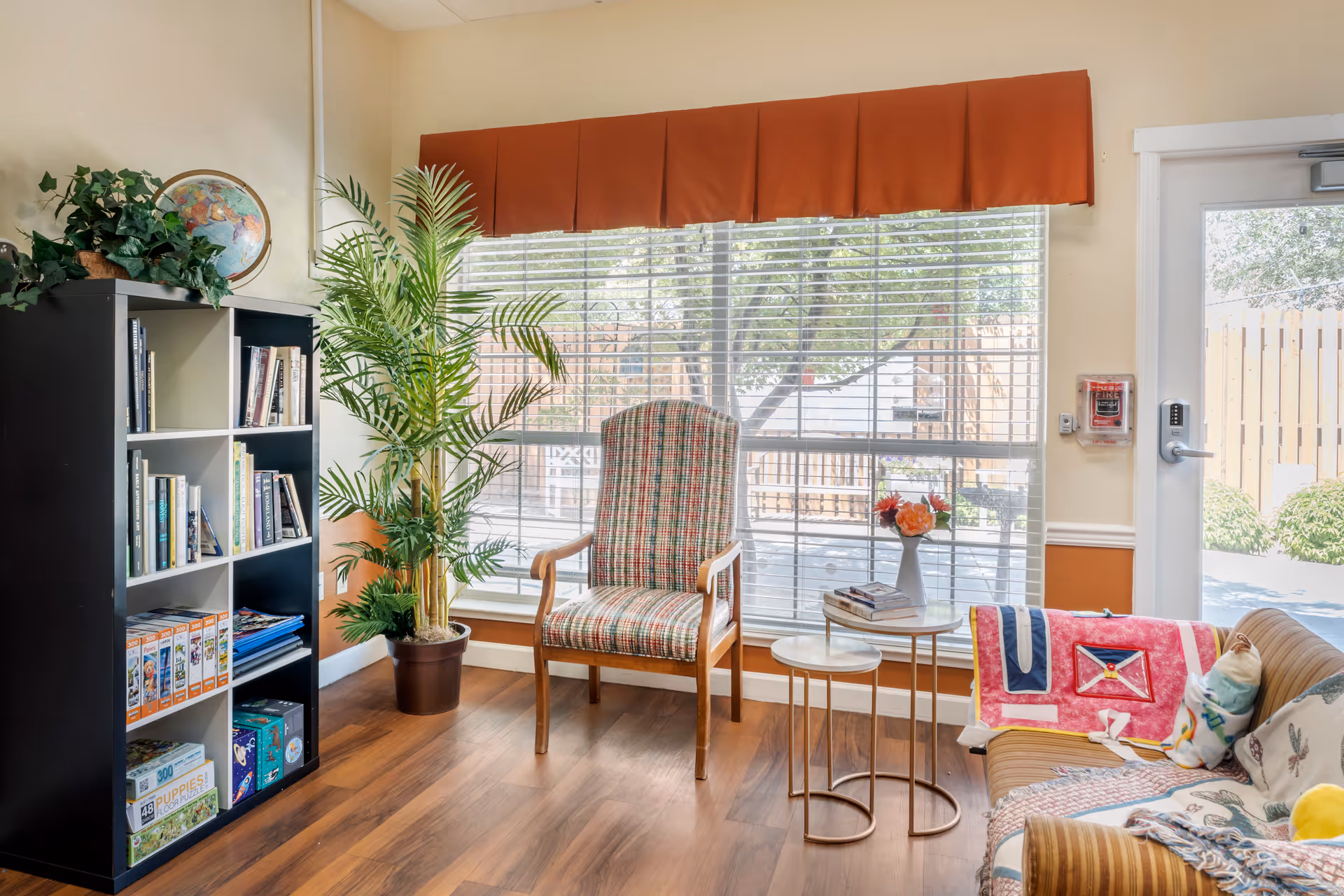 A cozy sunlit common room with a striped armchair, couch with colorful throws, small nesting tables, a bookshelf, and a large window with blinds.