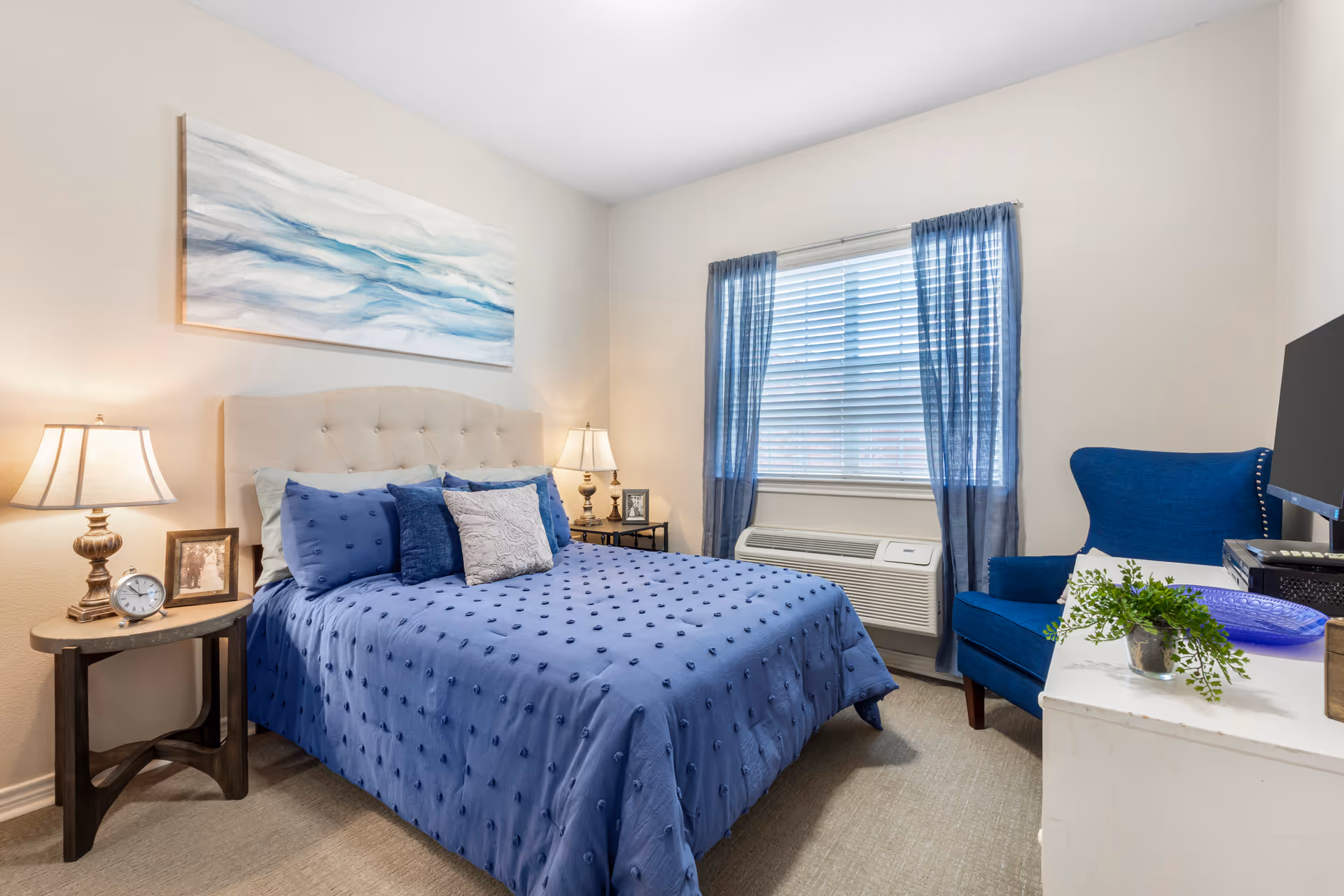 A cozy bedroom with a bed covered in a blue textured comforter and multiple pillows. There are two bedside tables with lamps and framed photos. A large window with blue curtains lets in natural light. A blue armchair is positioned next to a white dresser with a small green plant and a blue decorative plate. A painting with blue and white abstract waves hangs above the bed.