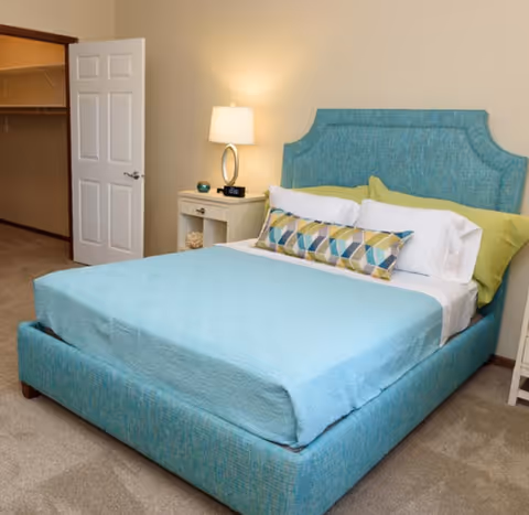 A neatly made bed with a turquoise upholstered headboard and matching bed frame, light blue bedspread, white and green pillows, and a decorative geometric-patterned pillow. Next to the bed is a white nightstand with a lamp, a small clock, and decorative items. An open door reveals a walk-in closet with shelving and hanging rods. The room has beige walls and carpeted flooring.