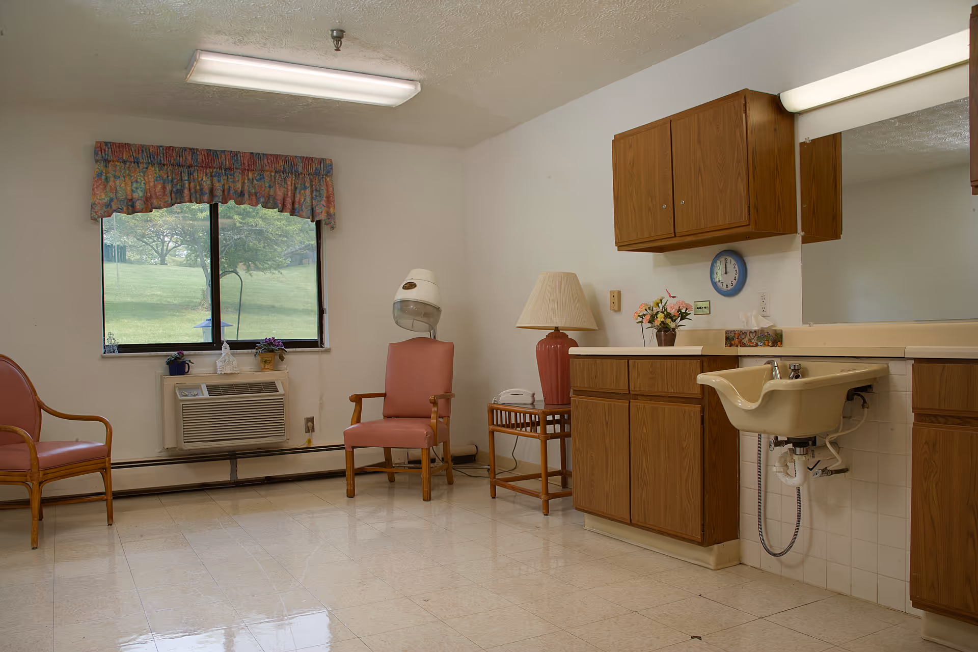 A room with two pink cushioned chairs, a hair dryer hood, a window with floral valance showing a green outdoor area, a wall-mounted air conditioner, a wooden cabinet with a countertop, a beige sink, a table lamp, a small side table with a telephone, and a large mirror above the sink and countertop.