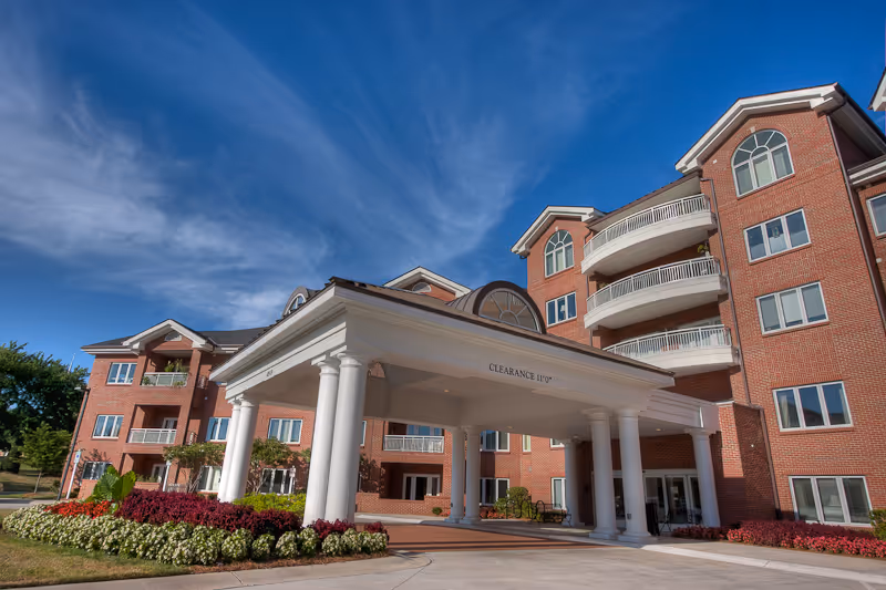 Exterior view of a multi-story brick senior living facility with white columns supporting a covered entrance. The building has balconies and large windows, with landscaped flower beds and a clear blue sky in the background.