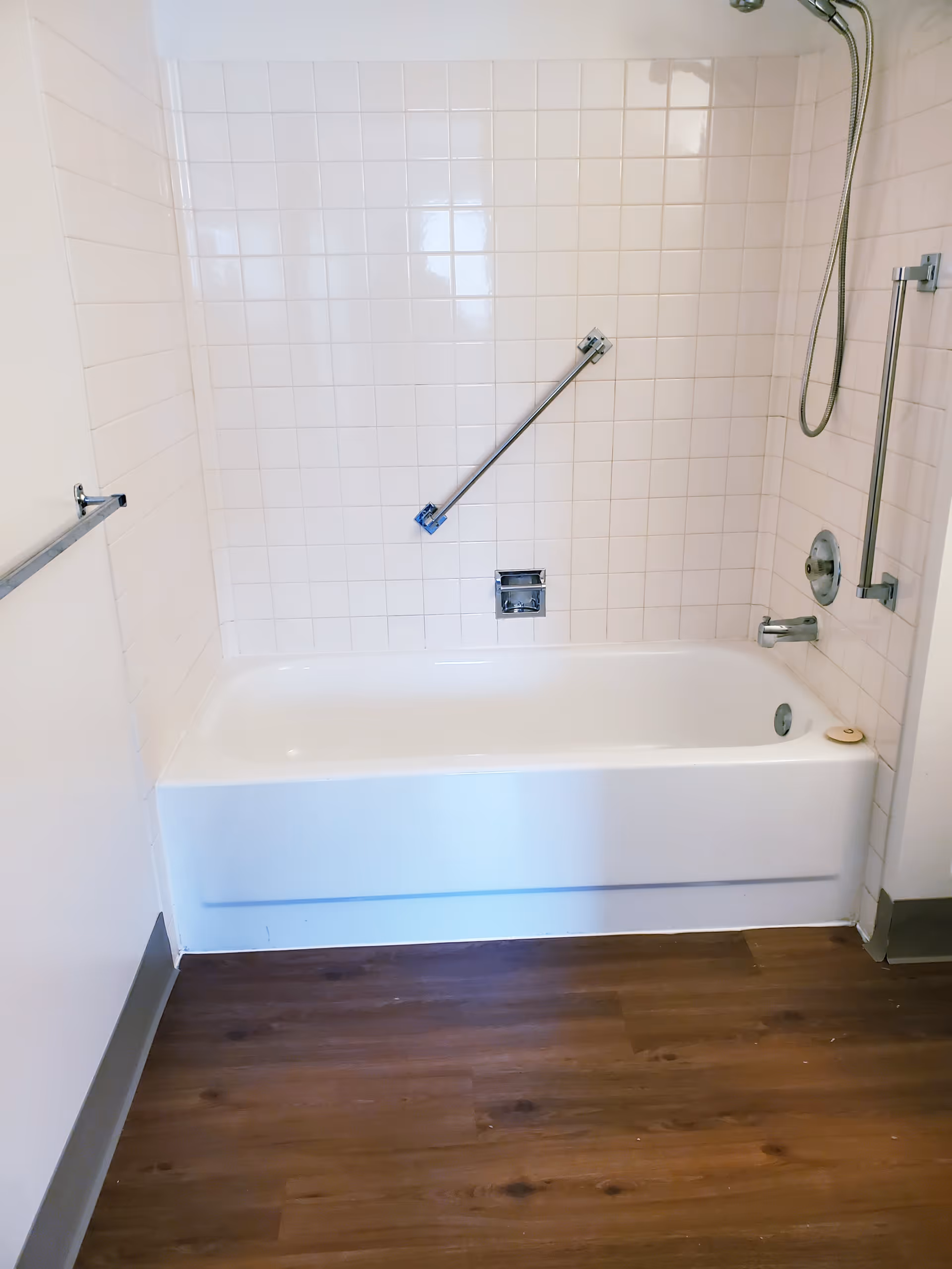 A clean bathroom with a white bathtub and white tiled walls. There is a metal grab bar installed diagonally on the wall above the bathtub, a showerhead with a flexible hose, and a faucet. The floor has wood-like vinyl flooring.