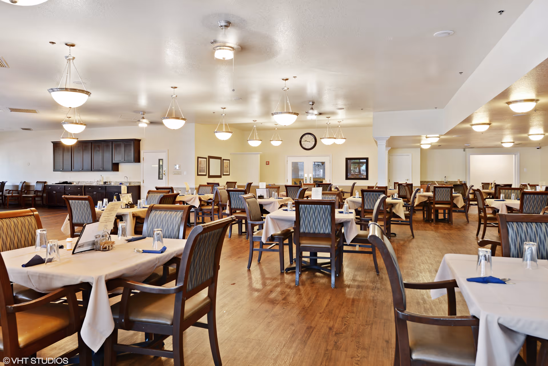 A spacious dining room with multiple tables covered in beige tablecloths, each set with glasses, napkins, and menus. The room features wooden chairs with patterned upholstery, wooden flooring, and several hanging light fixtures. There is a clock on the far wall above double doors, and cabinets along the left wall.