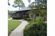 A single-story building with a sloped roof surrounded by green shrubs and trees. A curved concrete walkway leads to the entrance, and the sky is partly cloudy.