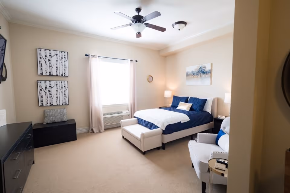 Well-lit bedroom with a blue-and-white bed, armchair, dresser, window and ceiling fan.