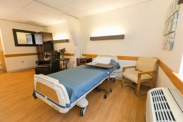 A patient room in a senior living facility featuring a hospital bed with a blue blanket, a beige armchair, a black recliner, a small table with flowers, a wall-mounted light, a telephone on a nightstand, and a heating/cooling unit near the window. The room has wood flooring and neutral-colored walls.