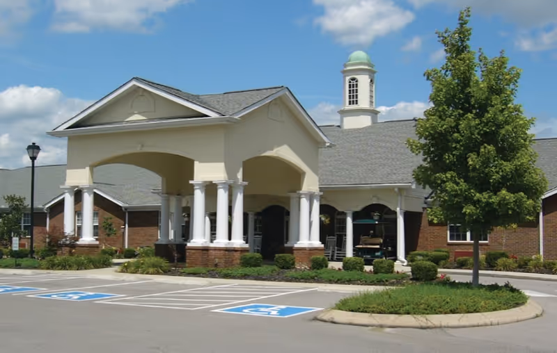 Exterior view of a senior living facility building with a covered entrance supported by white columns, a cupola on the roof, landscaped greenery, and designated handicap parking spaces in front.