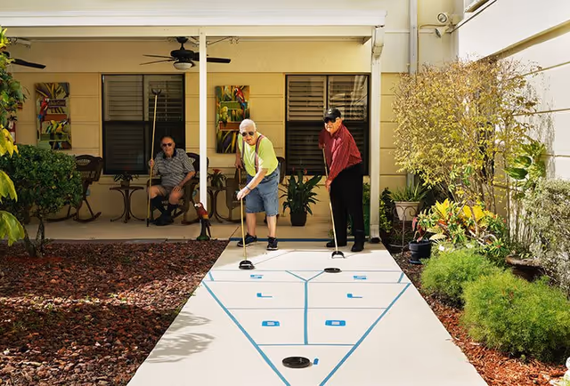 Two elderly men playing shuffleboard on an outdoor court at a senior living facility, with one man preparing to shoot and the other watching. Another elderly man is seated in the background on a covered patio area with chairs and plants around.