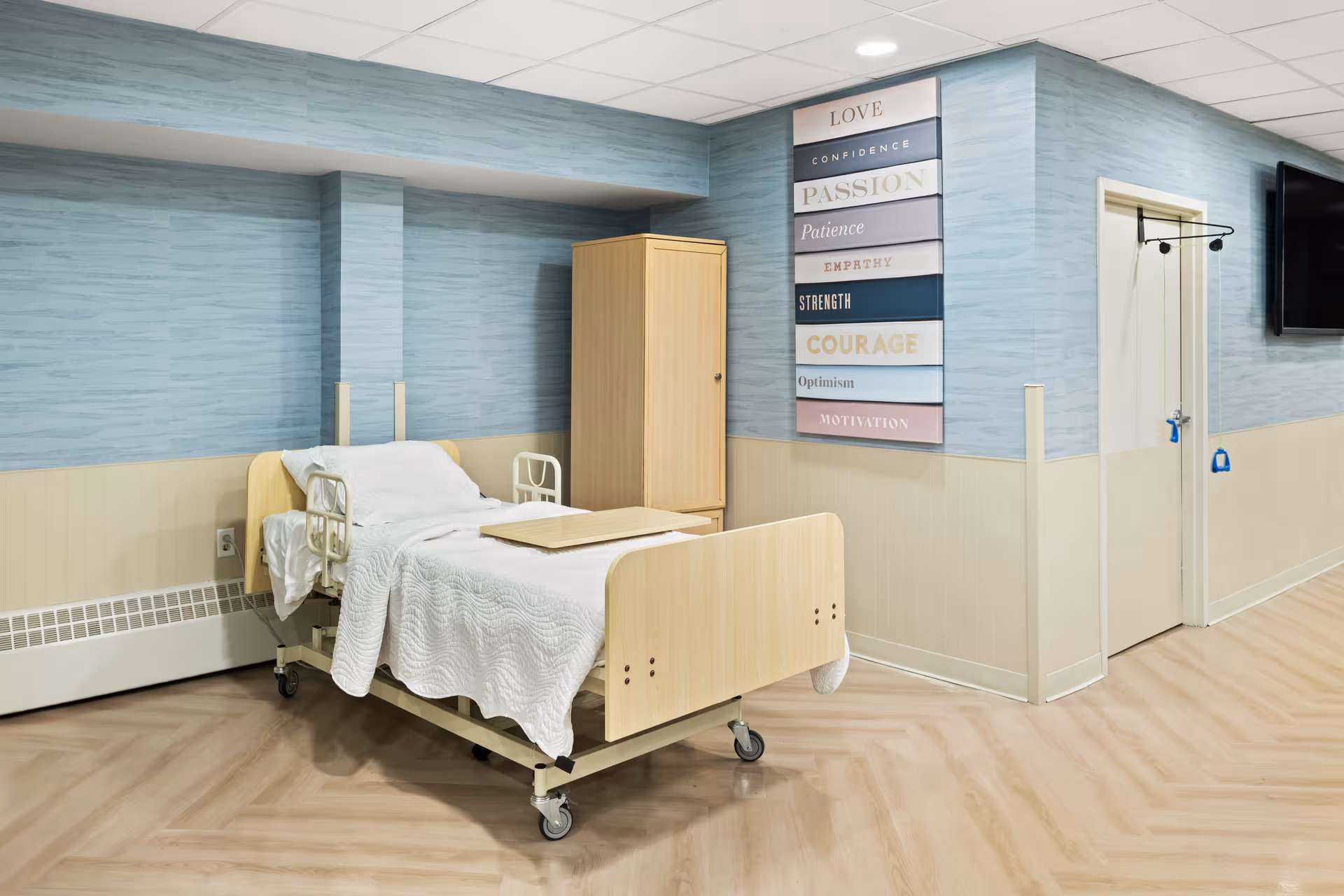 A hospital-style bed with white bedding and a wooden tray on top is positioned in a room with light blue and beige walls. There is a wooden cabinet next to the bed, a wall-mounted TV on the right, and a motivational wall art with words like Love, Confidence, Passion, Patience, Empathy, Strength, Courage, Optimism, and Motivation. The floor has a light wood pattern.