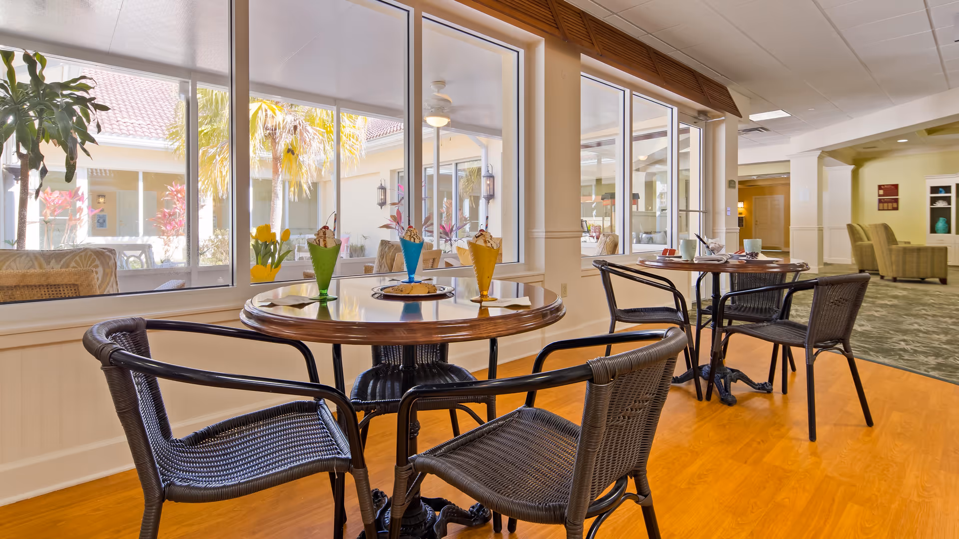 A bright indoor dining area with two round wooden tables, each surrounded by four black wicker chairs. On one table, there are three colorful ice cream sundaes and a plate with cookies. Large windows provide a view of an outdoor courtyard with palm trees and plants. The floor is wooden, and the background shows a hallway with carpet and additional seating.
