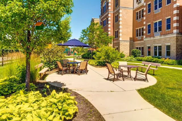 Outdoor patio area at Montclair Assisted Living and Memory Care with round tables and chairs, one table shaded by a blue umbrella, surrounded by green trees, bushes, and a well-maintained lawn next to a multi-story brick building under a clear blue sky.