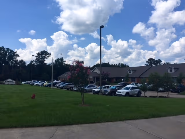 Exterior view of Copiah Living Center showing a single-story building with a parking lot filled with cars in front. There is a grassy area with a small tree and a fire hydrant in the foreground under a partly cloudy sky.