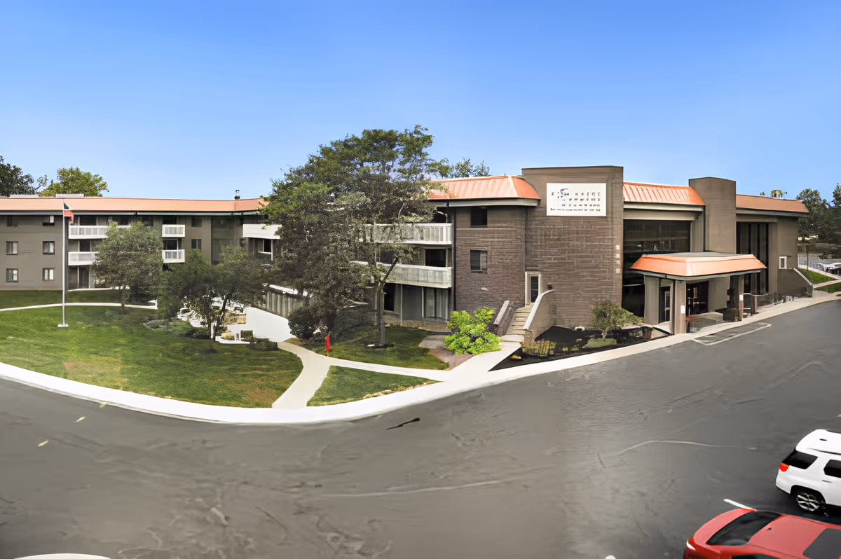 Exterior view of King's Reserve College Park senior living facility showing a multi-story building with a red roof, surrounded by trees and a well-maintained lawn with walkways. Several parked cars are visible in the foreground under a clear blue sky.