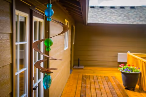 An outdoor wooden deck beside a tan building features a hanging bead ornament and potted flowers.