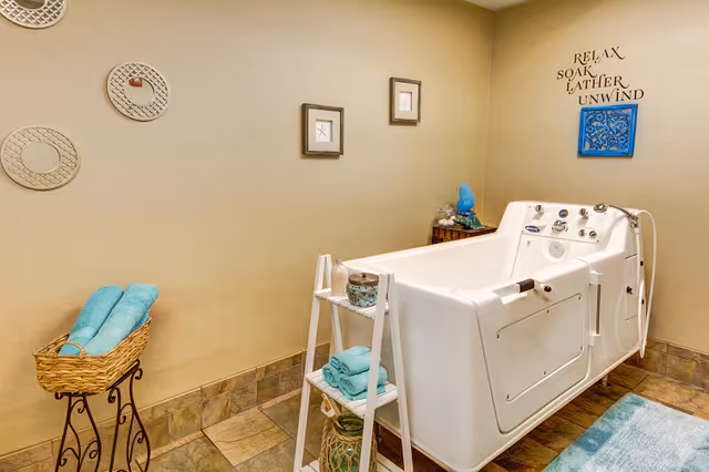 A walk-in bathtub in a bathroom with beige walls and tiled floor. The bathtub has multiple controls and a handheld shower head. There is a small white shelf next to the tub holding folded turquoise towels and decorative items. A wicker basket with turquoise towels is on a stand to the left. Wall decor includes round mirrors, framed pictures, and a blue decorative piece with the words 'RELAX SOAK LATHER UNWIND' above the bathtub.