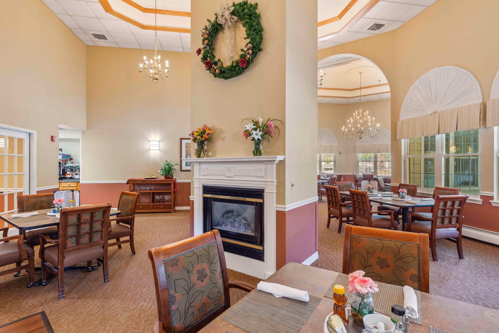A spacious dining room in a senior living facility with multiple tables and chairs arranged neatly. The room features high ceilings with chandeliers, large windows with decorative curtains, and a central fireplace adorned with a wreath and flower vases. The walls are painted in warm beige and maroon tones, creating a cozy and inviting atmosphere.