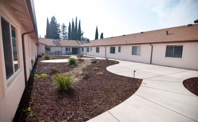 Outdoor courtyard area of Hospitality House Assisted Living & Memory Care with a curved concrete walkway, landscaped garden beds with mulch and small plants, and single-story building walls with windows surrounding the courtyard.
