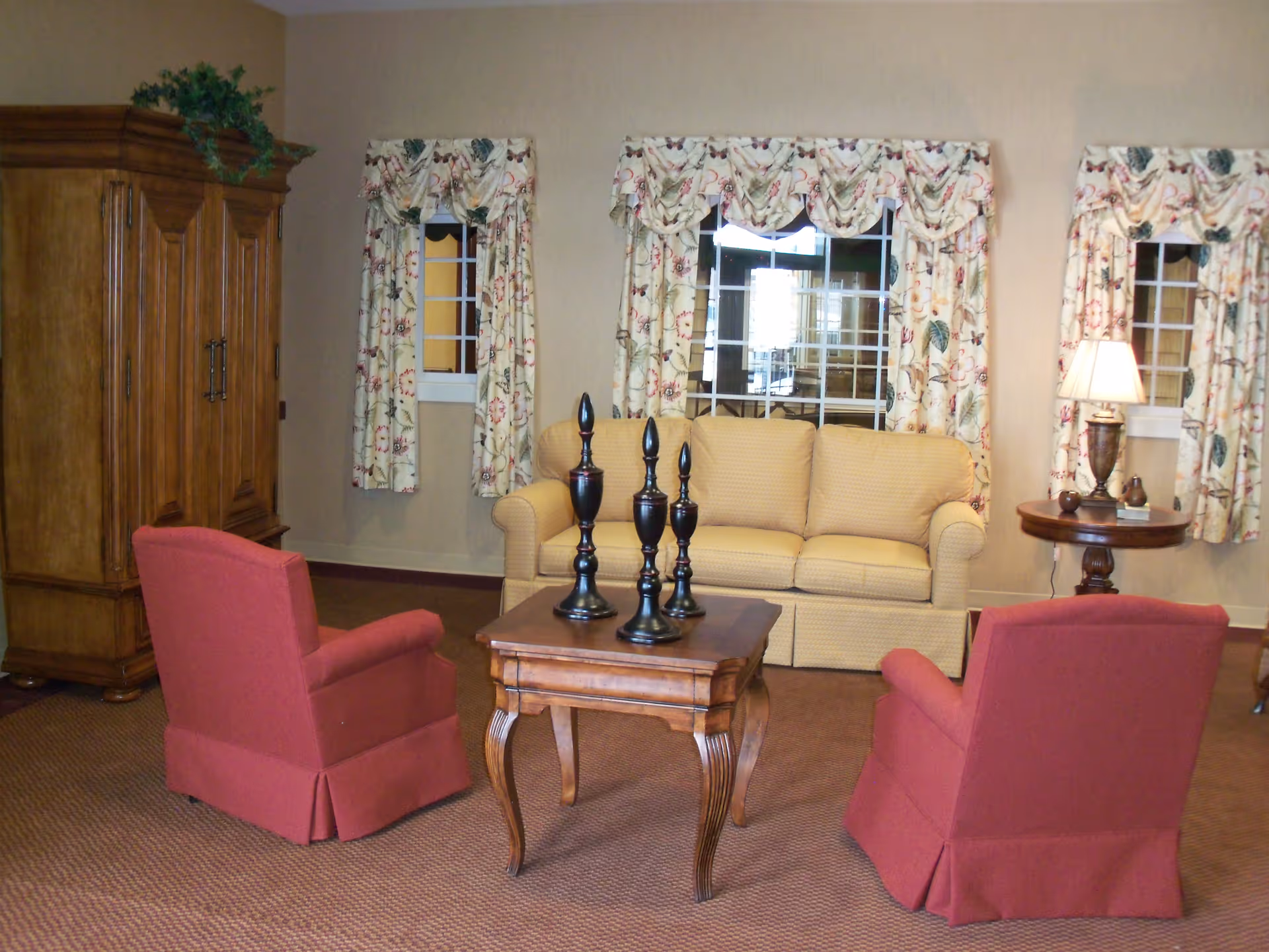 A cozy living room with a yellow sofa, two red armchairs, a wooden coffee table with three decorative black candle holders, floral curtains on three windows, a wooden armoire with a plant on top, and a round side table with a lamp and small decorative items.