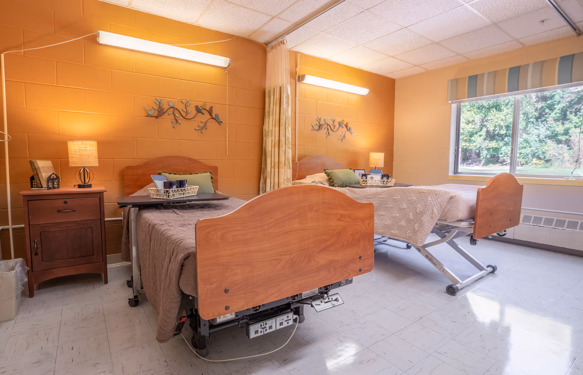 A bright room with two hospital-style beds separated by a curtain. Each bed has a wooden headboard and footboard, a beige quilt, and a green pillow. There are bedside tables with lamps and decorative items next to each bed. A large window with a striped valance lets in natural light, and the walls are painted orange and beige.