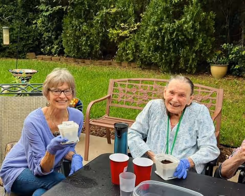 Two elderly women sitting outdoors at a black table, smiling and holding small containers with soil, wearing gloves. Behind them is green grass, bushes, and outdoor furniture including a bench and a chair.