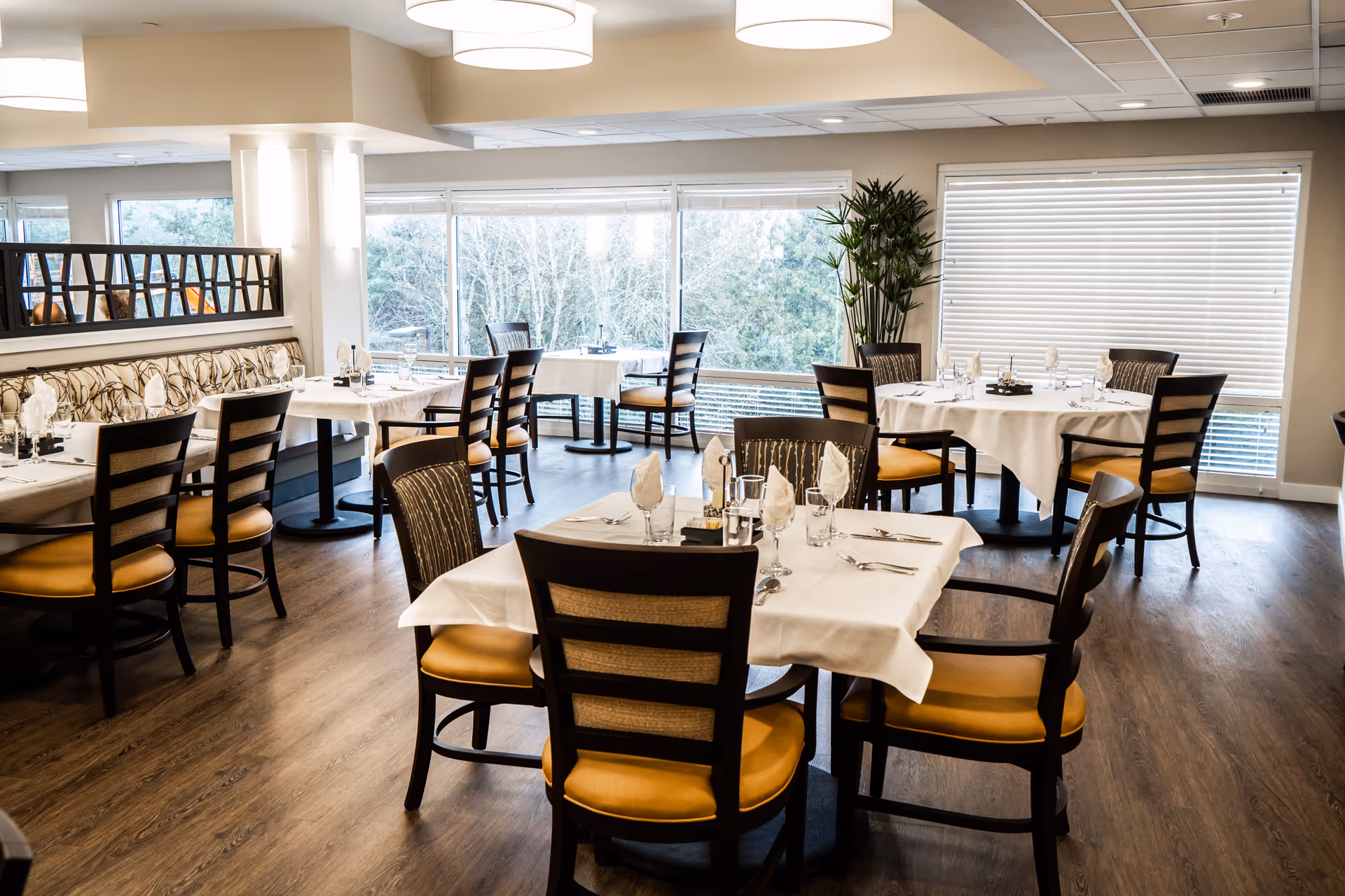 Dining room with multiple tables set with white tablecloths and chairs, large windows showing trees outside.