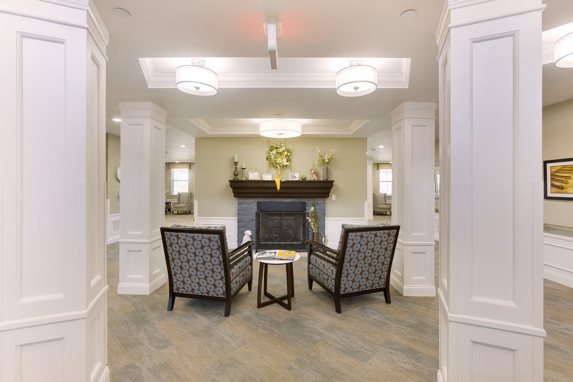 A cozy seating area in a senior living facility featuring two patterned armchairs facing a small round table with magazines on it, positioned in front of a fireplace with a dark wooden mantel decorated with flowers, candles, and framed photos. The space is framed by white columns and has soft lighting from ceiling fixtures.