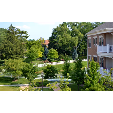View of a landscaped outdoor area with green trees, shrubs, and a white fence next to a multi-story brick building with balconies at Maple Knoll Village.