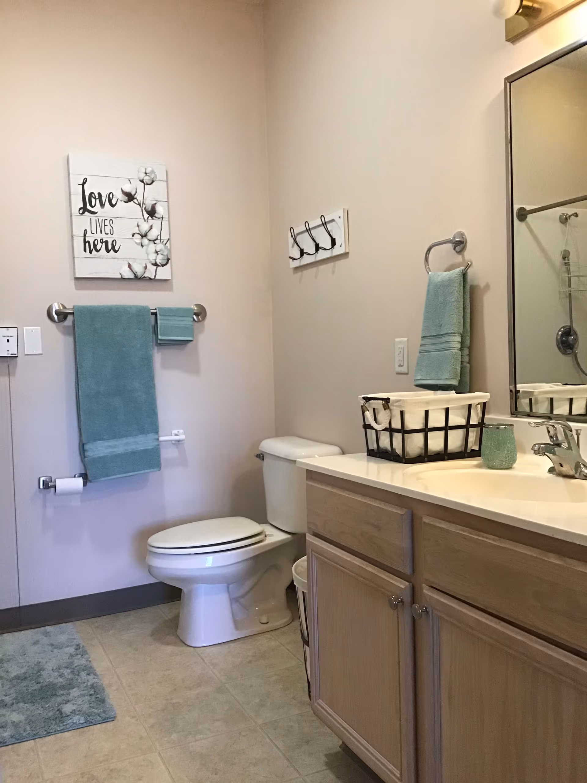 A clean bathroom featuring a toilet, a wooden vanity with a sink, a large mirror, and teal towels hanging on towel racks. There is a decorative wall sign that reads 'Love lives here' and a basket with rolled towels on the vanity countertop.