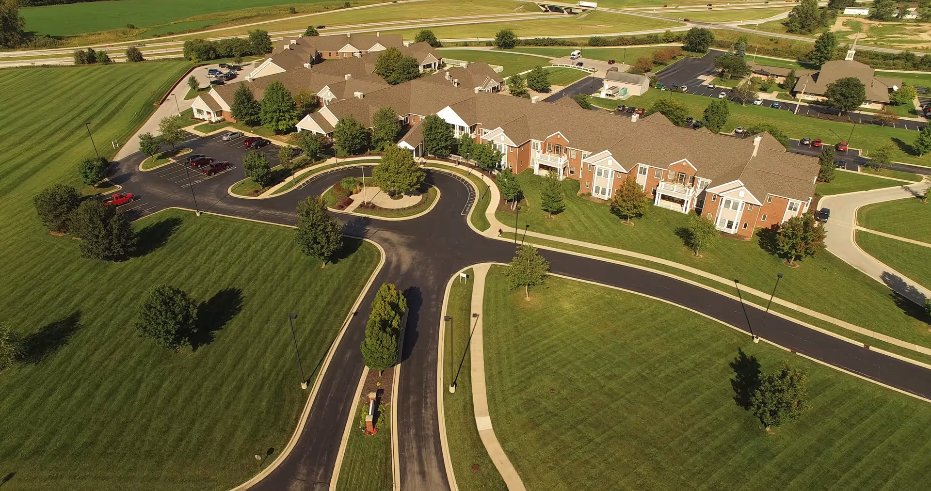 Aerial view of Heritage Pointe of Huntington, showing a large senior living facility surrounded by well-maintained green lawns, trees, and paved roads with parking areas. The building has multiple sections with brown roofs and light-colored walls, and there are several cars parked near the entrance.