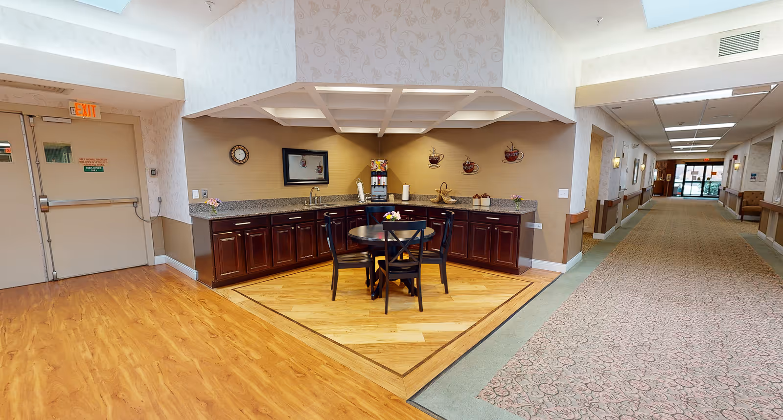 A bright interior hallway area featuring a small kitchenette with dark cabinets and a round table with chairs on a wood inset, leading down a long carpeted corridor.