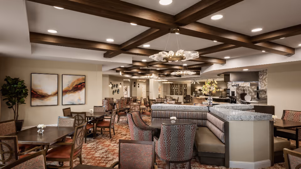 A spacious dining room with multiple tables and chairs arranged neatly. The ceiling features wooden beams with modern circular light fixtures. There are decorative paintings on the wall and a plant in the corner. The room has a warm and inviting atmosphere with a mix of upholstered chairs and booth seating.