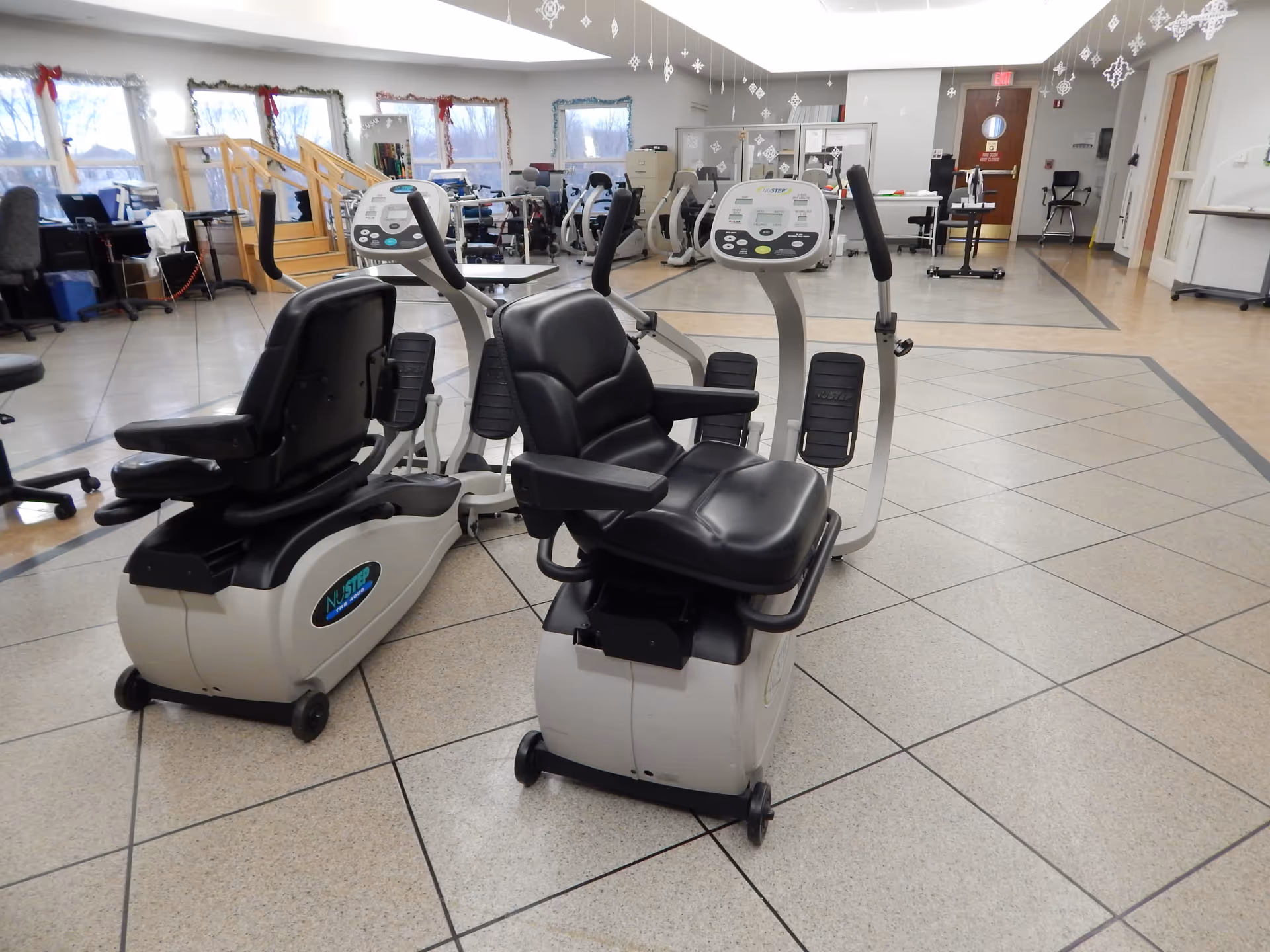 Indoor exercise room with two NuStep recumbent cross trainers in the foreground, several other exercise machines and equipment in the background, and windows decorated with holiday garlands and red bows.