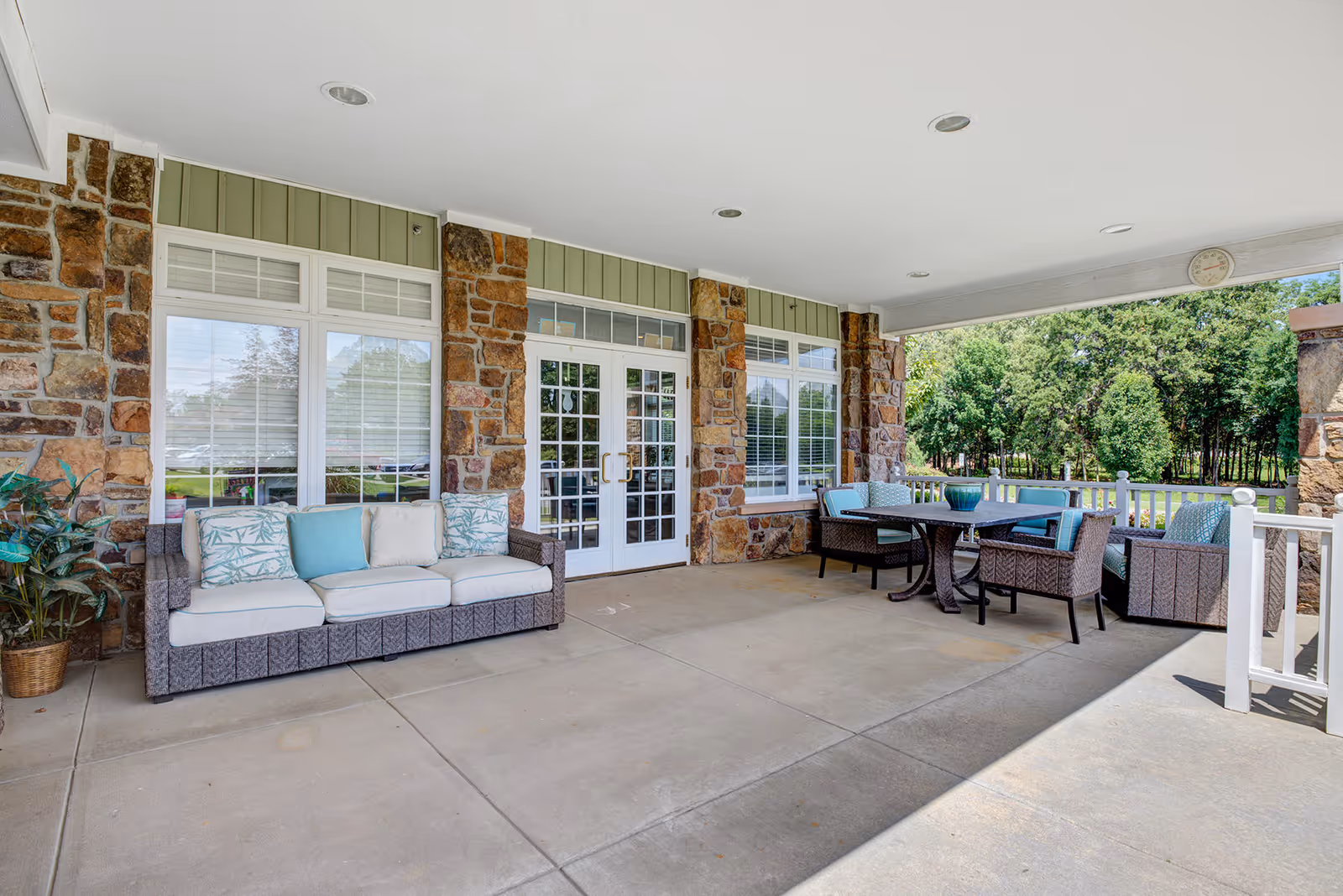 Covered outdoor patio area with stone pillars and white-framed windows and doors. There is a wicker sofa with cushions and pillows on the left, and a wicker dining set with chairs and a table on the right. Green trees and grass are visible beyond the patio.