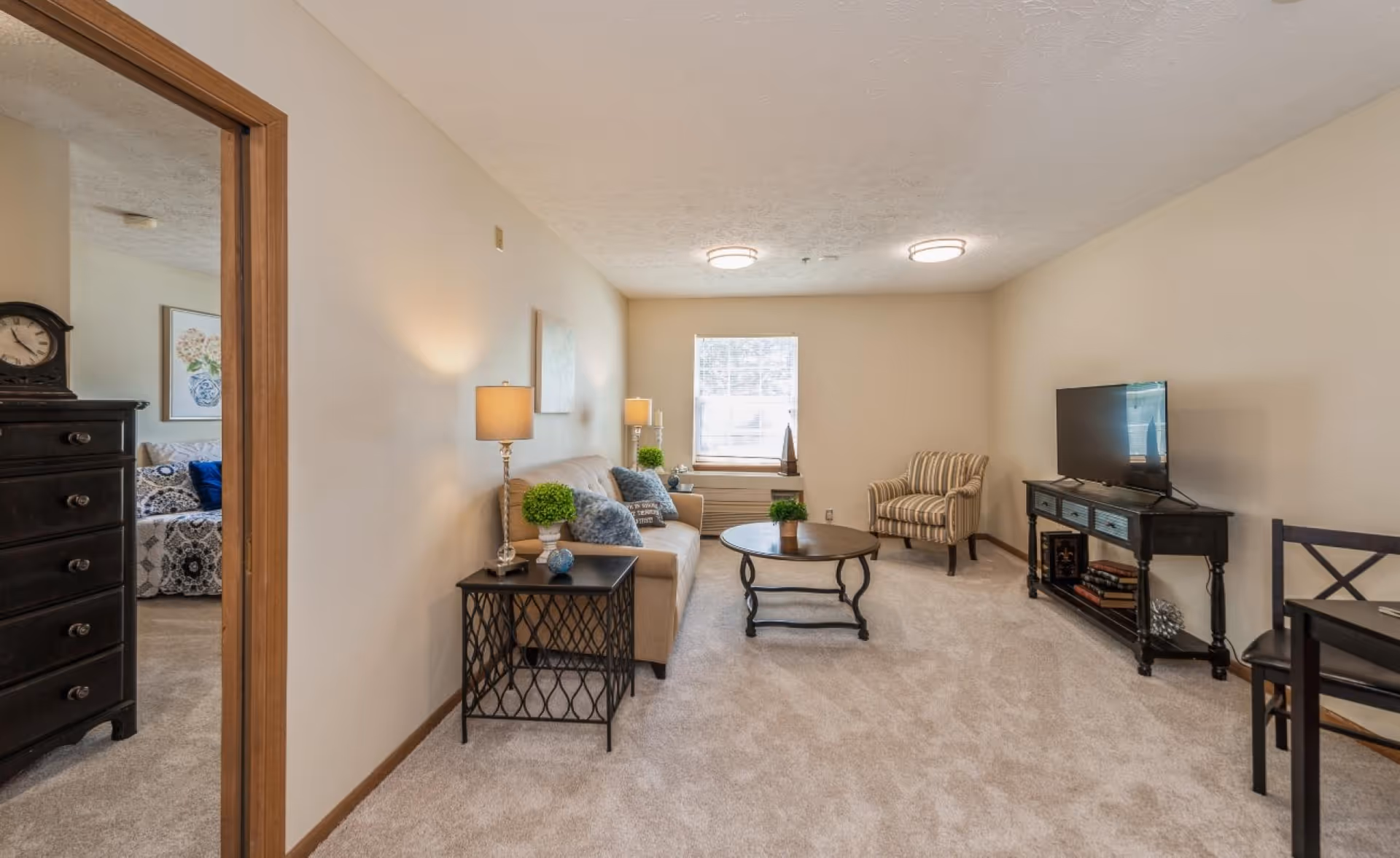 A cozy living room with beige walls and carpet. The room features a beige sofa with decorative pillows, a round wooden coffee table with a small plant, and a striped armchair near a window with blinds. A black TV stand holds a flat-screen TV and some books. To the left, an open doorway leads to a bedroom with a black dresser and a bed with patterned bedding. The room is softly lit by two ceiling lights and a table lamp on a black side table next to the sofa.
