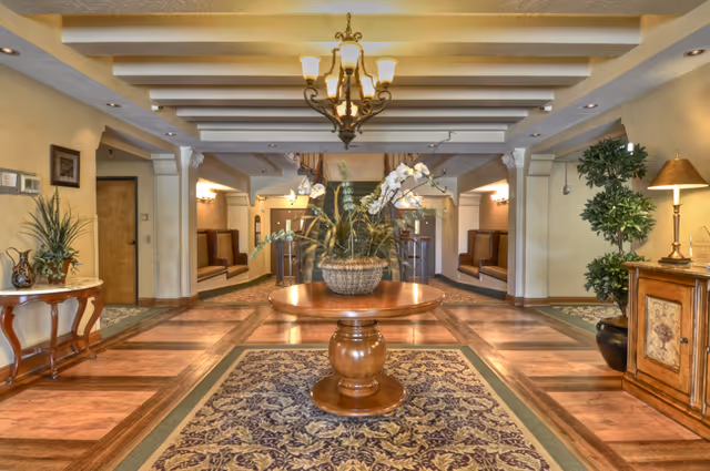 Interior view of a senior living facility lobby with a round wooden table in the center holding a large floral arrangement. The floor features a patterned rug and wood flooring. There are built-in seating booths along the walls, decorative plants, a chandelier hanging from the ceiling, and warm lighting throughout the space.