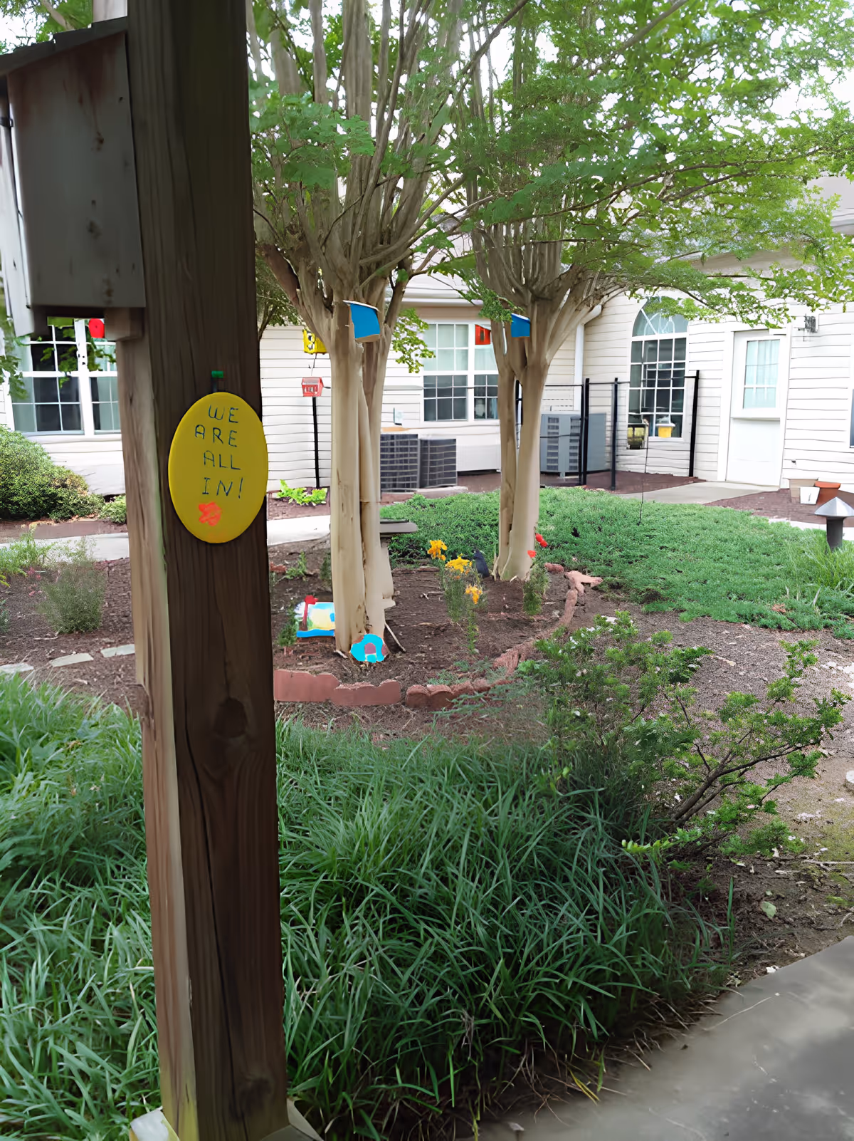 Courtyard garden with trees, shrubs, and a wooden post displaying a yellow sign that reads "WE ARE ALL IN!" in front of a senior living building.