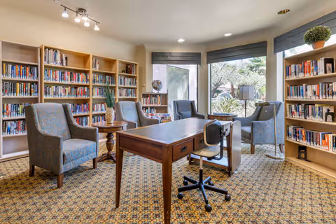 A cozy library room with bookshelves filled with books along the walls, several upholstered armchairs, a wooden table with a rolling office chair, and large windows letting in natural light with a view of greenery outside.