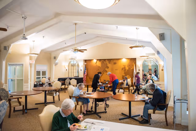 A spacious dining room in Solheim Senior Community with several elderly residents seated at round tables eating and engaging in conversation. The room has high vaulted ceilings with ceiling fans and pendant lights. There is a large wall decoration featuring a world map, and an American flag is visible in the background. The atmosphere is bright and welcoming.