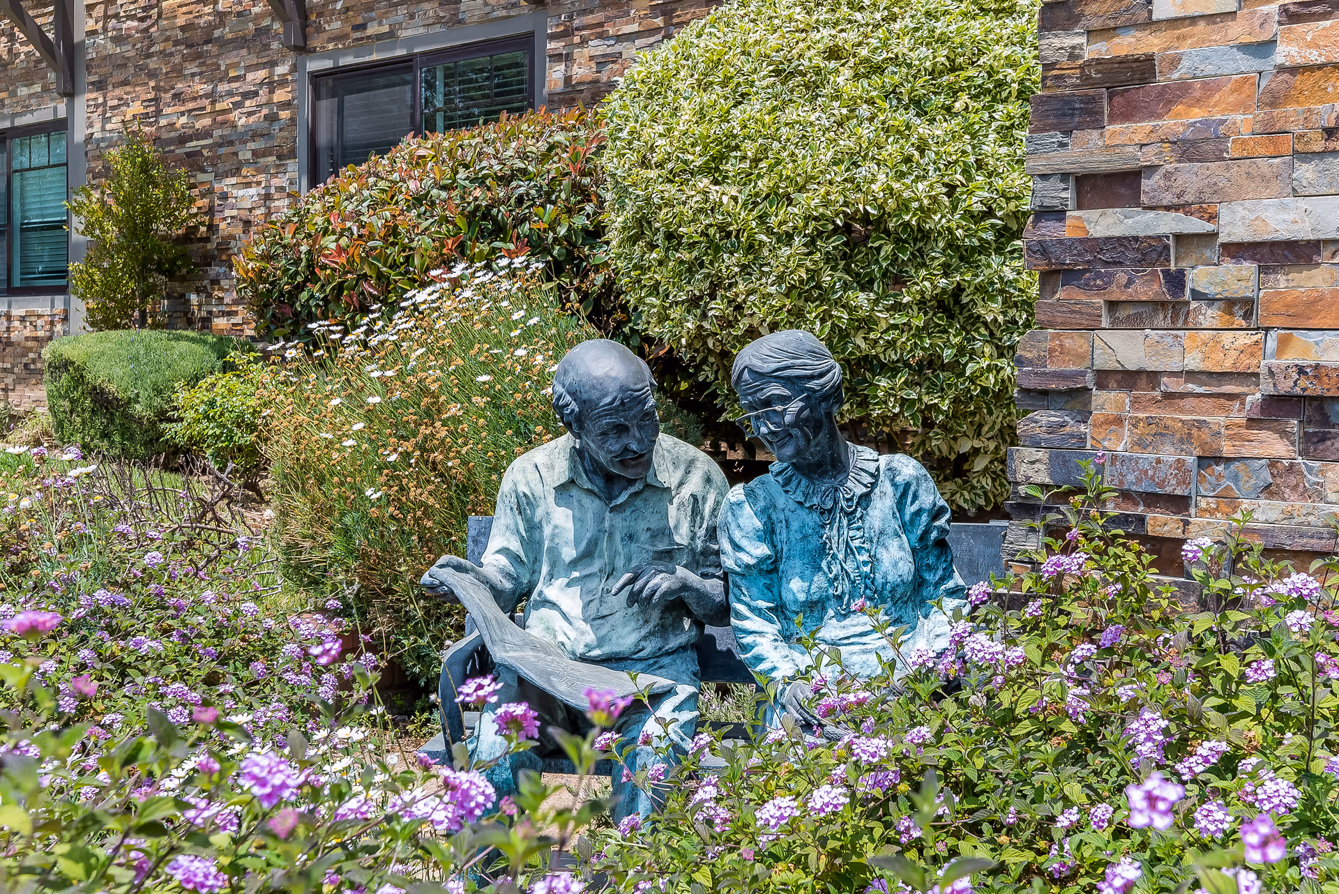 A garden area with colorful flowers and bushes surrounding a bronze statue of an elderly man and woman sitting on a bench, with a stone building wall in the background.