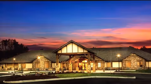 Exterior view of a single-story senior living facility building at sunset with warm lights illuminating the entrance and surrounding area, featuring a peaked roof and stone facade.