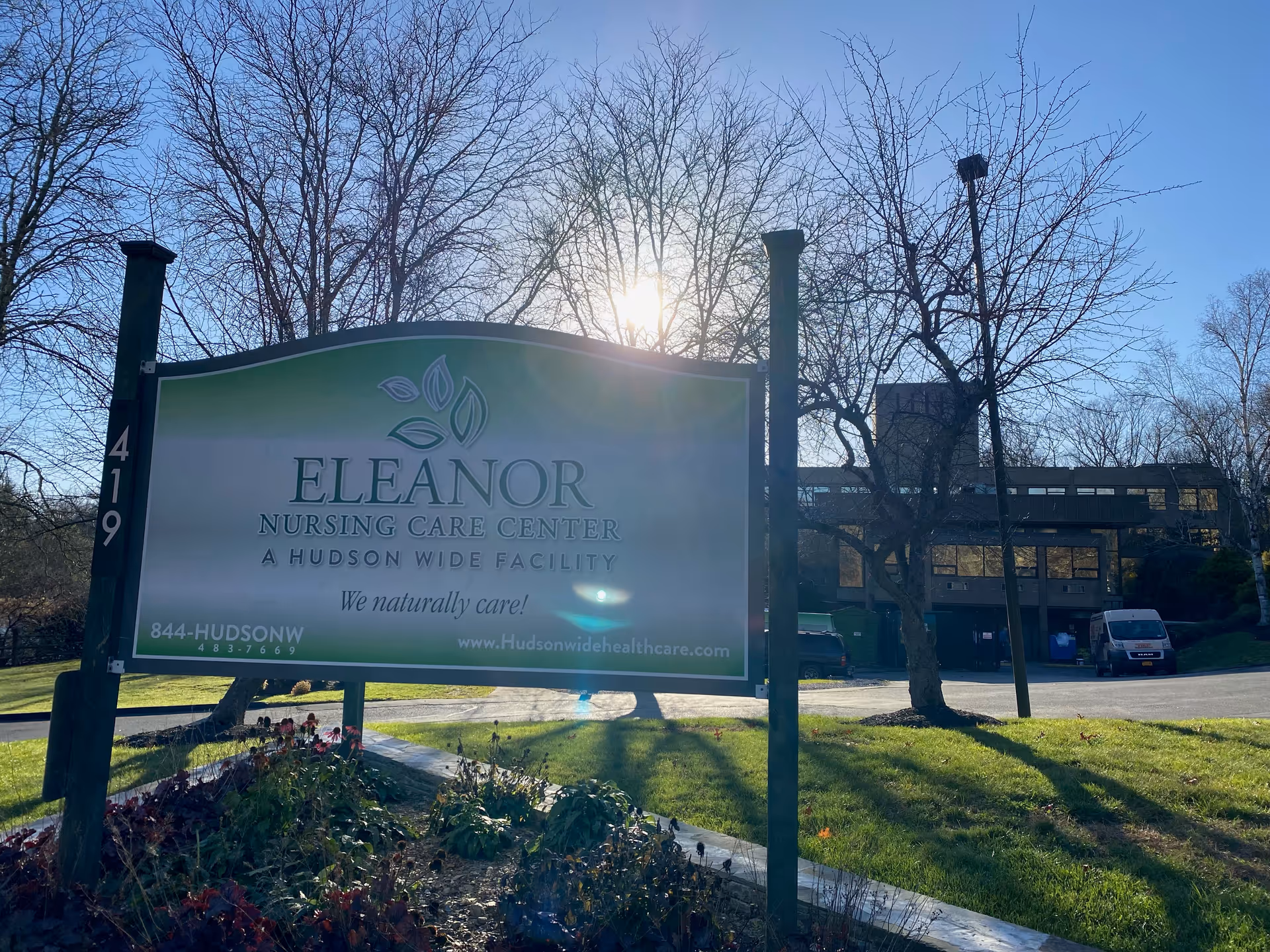 Outdoor view of the Eleanor Nursing Care Center sign with the building and leafless trees in the background under a clear blue sky with the sun shining through the trees.