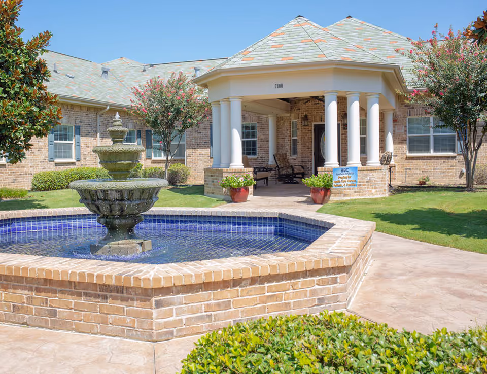 Front entrance of a brick senior living facility with a tiled fountain in the courtyard and a columned covered porch.
