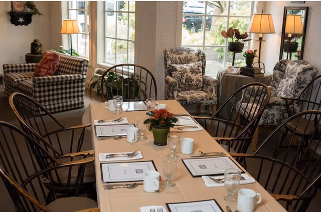 A cozy dining area with a rectangular table set for six, featuring menus, white mugs, glasses, and silverware. The table has a beige tablecloth and a small potted plant centerpiece. Surrounding the table are wooden chairs with curved backs. In the background, there are large windows letting in natural light, two floral-patterned armchairs, a checkered sofa with a red pillow, two table lamps, and some decorative plants.